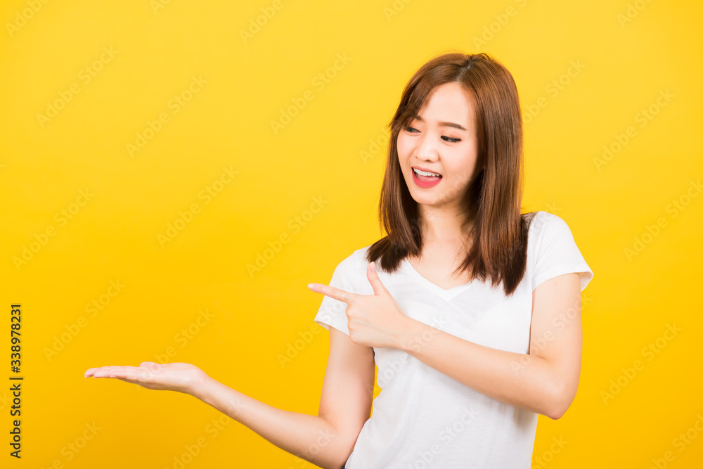 Asian happy portrait beautiful cute young woman teen standing wear t-shirt holding something on palm and point away side looking to hand isolated, studio shot on yellow background with copy space Asian happy portrait beautiful cute young woman teen standing wear t-shirt holding something on palm and point away side looking to hand isolated, studio shot on yellow background with copy space