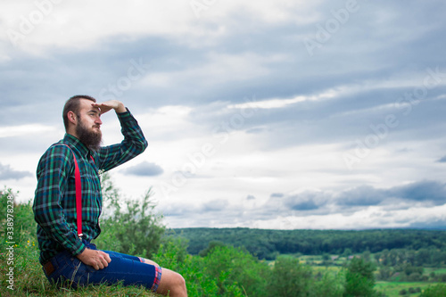 Handsome man hipster with beard on serious face in cloth shirt and suspenders sunny outdoor on mountain top against cloudy sky on natural. Tourism concept. Wonderful landscape.