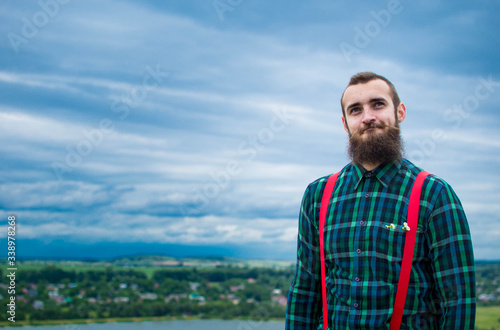 Handsome man hipster with beard on serious face in cloth shirt and suspenders sunny outdoor on mountain top against cloudy sky on natural. Tourism concept. Wonderful landscape.
