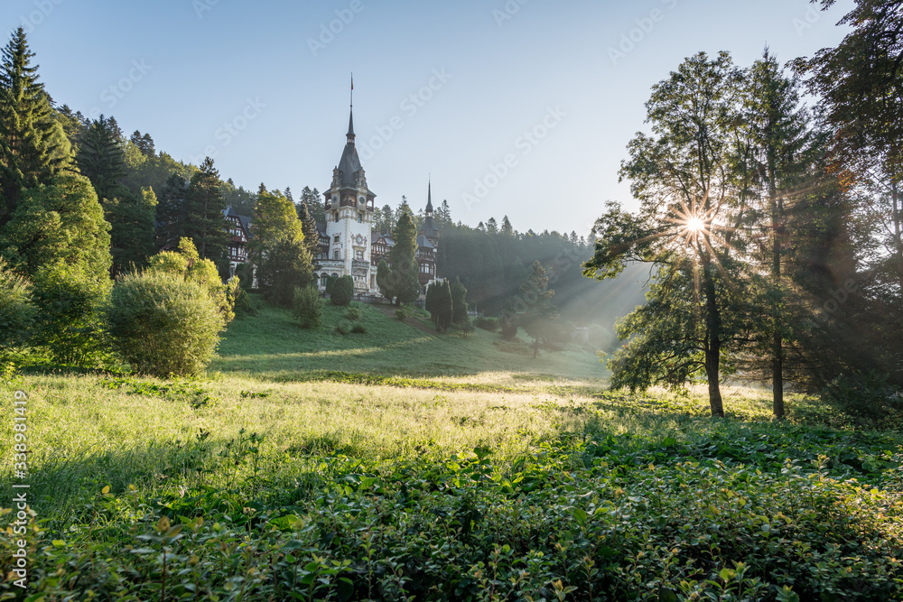 Peles Castle, famous residence of King Charles I in Sinaia, Romania. Peaceful summer landscape at sunrise of royal palace and park with mist and sun rays shining through the branches.