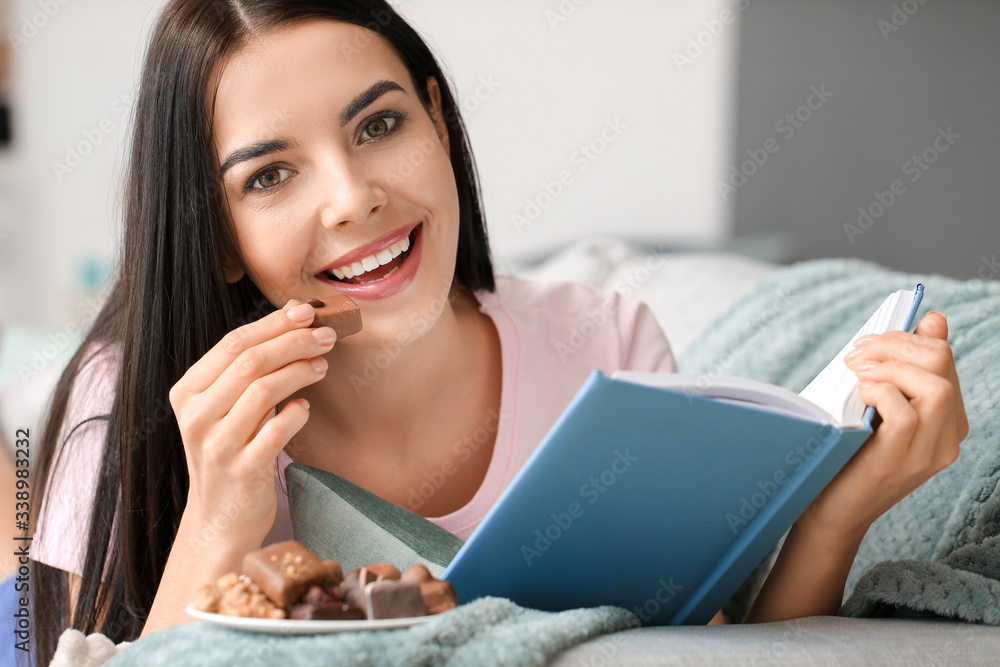 Beautiful young woman eating chocolate while reading book at home Beautiful young woman eating chocolate while reading book at home