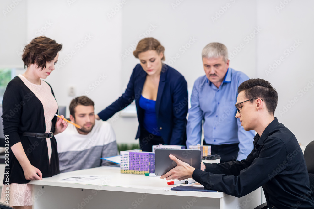 A young man showing a presentation to his colleagues at the meeting using his tablet and the models of houses A young man showing a presentation to his colleagues at the meeting using his tablet and the models of houses
