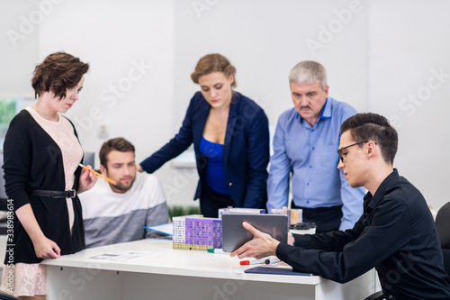 A young man showing a presentation to his colleagues at the meeting using his tablet and the models of houses