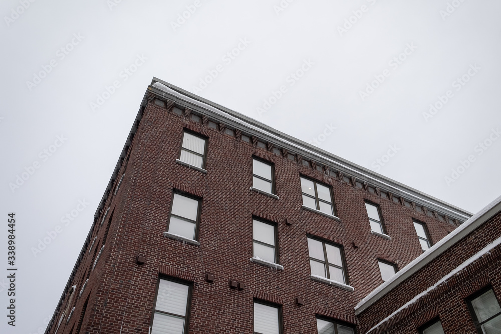 The top corner of a brick building with small single and double hung windows on three floors. The brown brick has a tan colored trim. The sky in the background is grey with clouds. 