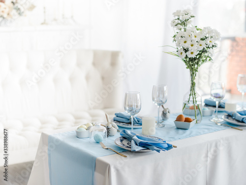 Decorative festive table setting with sky blue textile napkin and tablecloth, eggs, floral dish and golden cutlery, glass for wine. Happy easter concept.