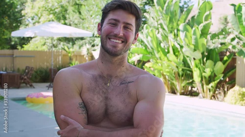 Portrait of smiling bare chested man standing by swimming pool with group of friends enjoying summer pool party together - shot in slow motion