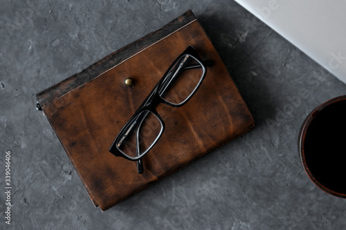 Brown leather notebook, pen, laptop and glasses on gray background