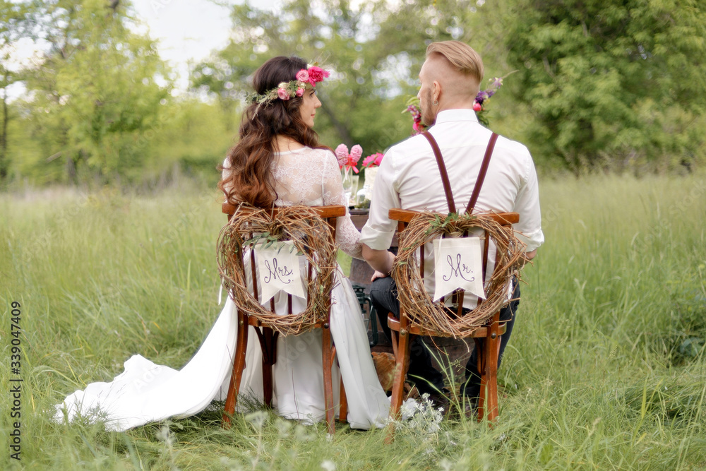 The bride in rustic style with a beautiful wreath on the head of flowers in a park in white dress with groom white hair in white shirt suspenders sit at a table with wine sweets and a wedding cake