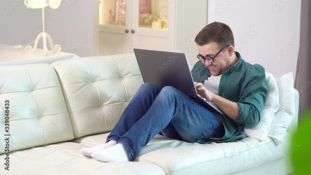 Funny male in glasses is typing on laptop on sofa