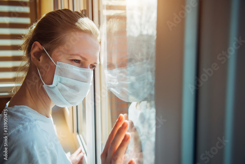 Sad woman in a protective medical mask looks out the window, close up. Self-isolation, quarantine, stay at home. Protective against Covid-19.