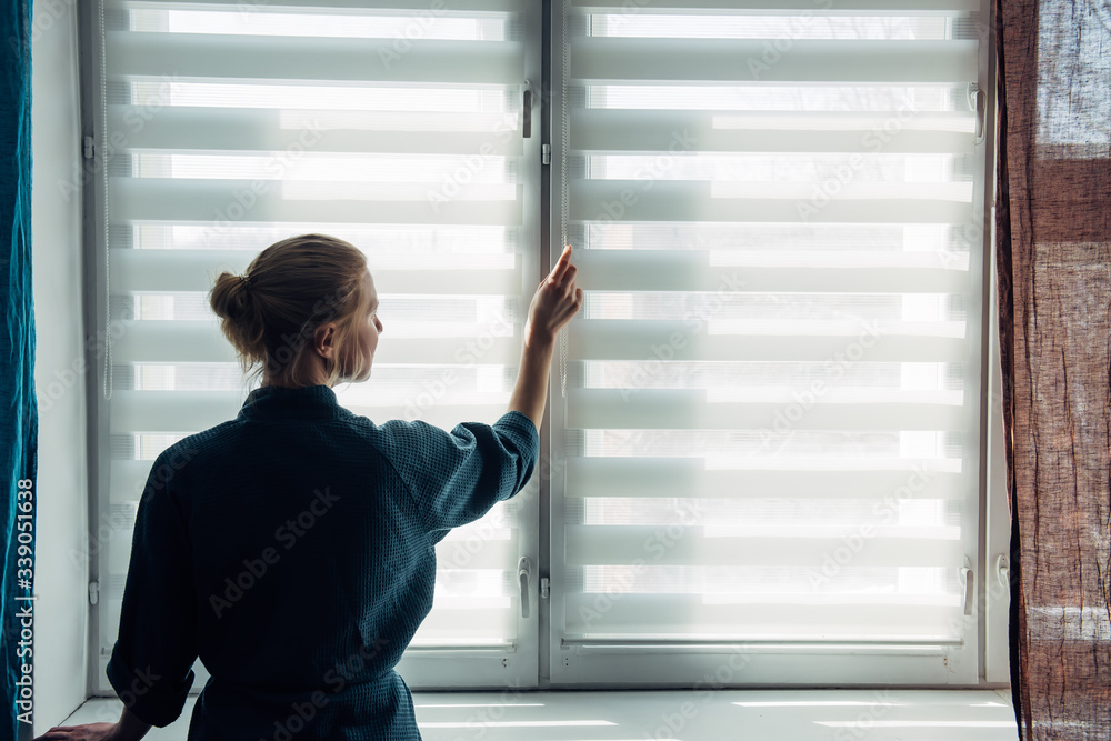 Woman's silhouette at window in the background light. Young woman in gown stands near window with blinds. Quarantine, self-isolation, stay home, self-preservation, coronavirus pandemic. Woman's silhouette at window in the background light. Young woman in gown stands near window with blinds. Quarantine, self-isolation, stay home, self-preservation, coronavirus pandemic.