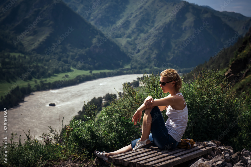 Young woman in white t-shirt and shorts sits on top of hill and admires the beautiful view of the mountains and river on snny summer day. Pretty blonde resting while hiking in the mountains.