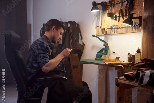 The leather master is working with natural leather at the desk. The room with wooden table and set of leather craft tools on it. Tools for craft production of leather goods on work desk.