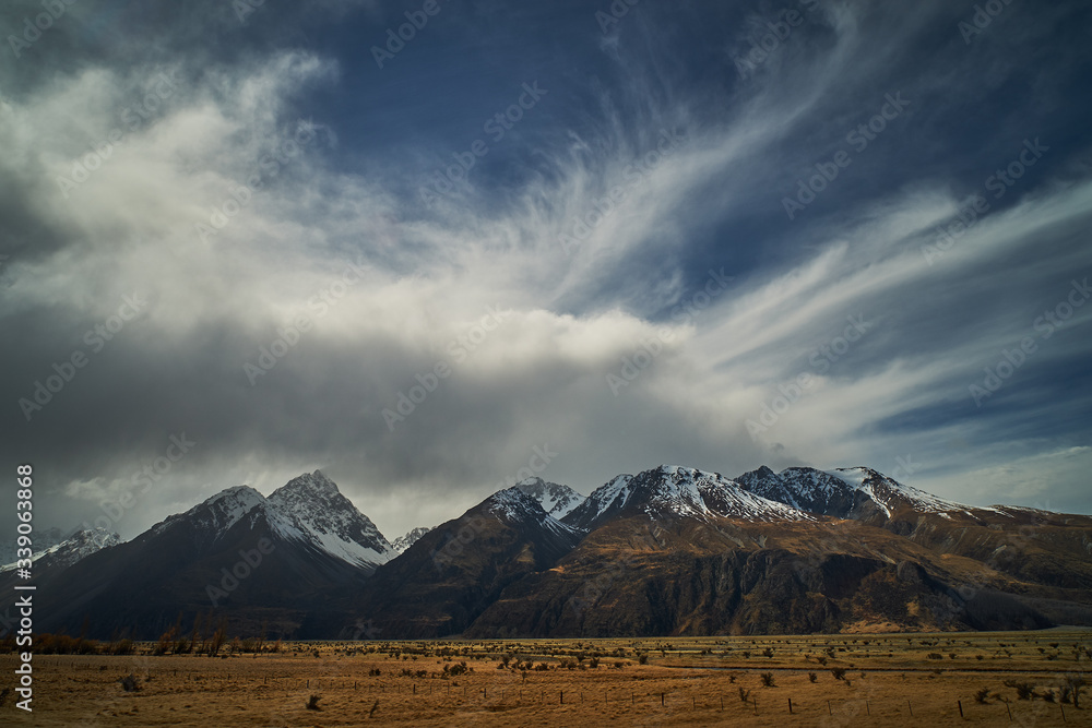 
Fields of brown alpine grass landscape with snow covered mountains in the background and clouds overhead at Mt Cook in New Zealand South Island
