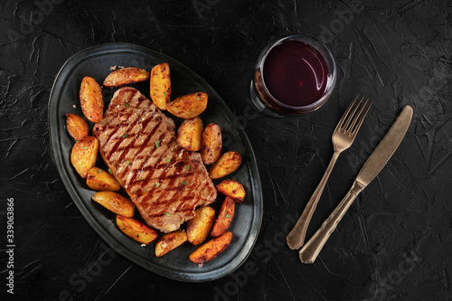Grilled beef steak with baked potato wedges, with a glass of wine, shot from the top on a black background