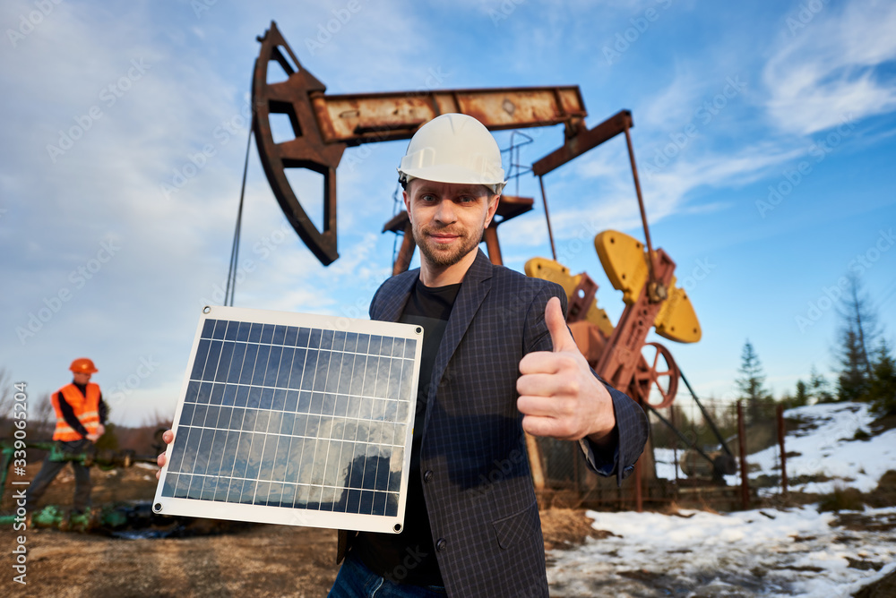 Portrait of smiling businessman in protective helmet holding portable solar panel and showing approval gesture. Oil man engineer working with pump jack on territory of oil field on background. Portrait of smiling businessman in protective helmet holding portable solar panel and showing approval gesture. Oil man engineer working with pump jack on territory of oil field on background.