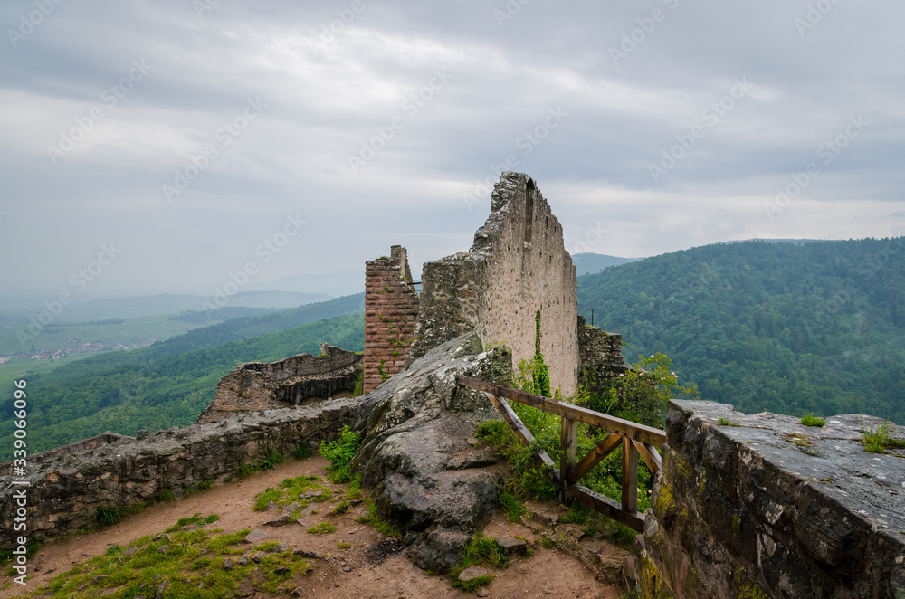 A ruined medieval castle tower on a hill with mountains in the background and cloudy sky above in France.