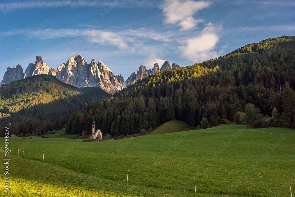 A tiny church surrounded by fields of grass and forest in the beautiful valley of Val di Funes with a view of The Dolomites Geisler Odle mountain peaks in the background