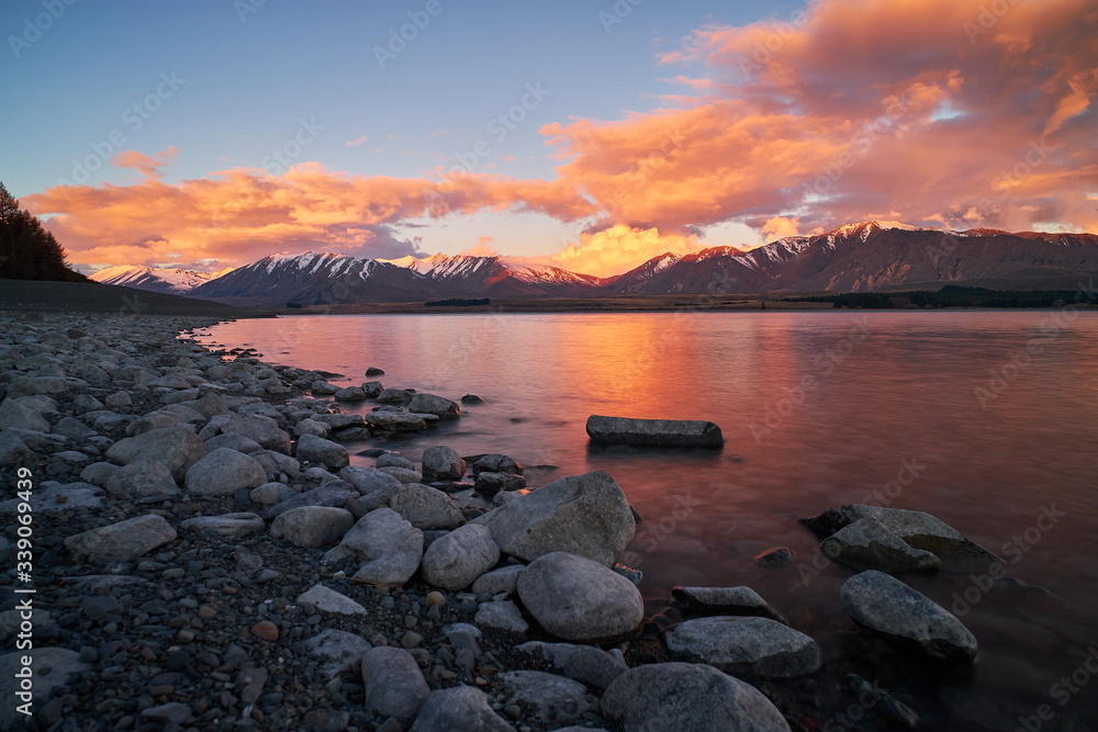 A beautiful colourful sunset with snow capped mountains from a rocky shore in Lake Tekapo in New Zealand South Island