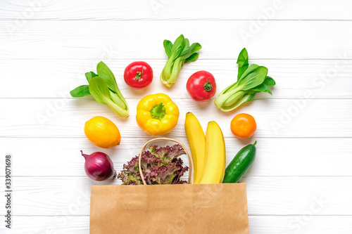 Full eco paper bag of different health food - yellow bell pepper, tomatoes, bananas, lettuce, green, cucumber, onions on white wooden background Top view Flat lay Grocery shopping