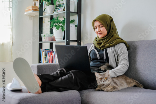 Young happy asian muslim female adult sitting at the sofa, working from home due to lockdown and quarantine from covid-19 virus outbreak, accompany by domestic cat