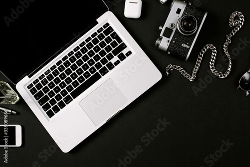 Stylish workspace of designer with laptop, vintage film camera, notebook, headphones, pen and smartphone on black desk. Flat lay, top view
