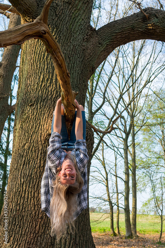 Junge Frau hängt lachend Kopfüber an einem Ast von einem dicken Baum. Ansicht von vorne. Standort: Deutschland, Nordrhein Westfalen, Hoxfeld photo