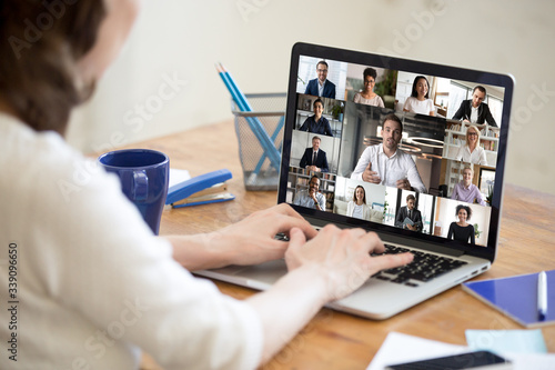 On laptop screen web cam view diverse age and ethnicity businesspeople taking part at group videocall, view over woman shoulder sit at desk working from home. Distant communication, modern app concept photo