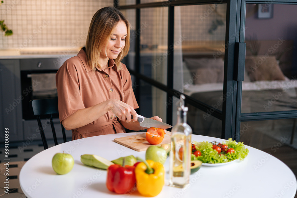 Happy smiling woman cutting fresh organic tomato with a knife for vegetable salad in modern kitchen. Smiling slim vegetarian female prepare healthy vegan dinner meal. Concept of healthy eating. Happy smiling woman cutting fresh organic tomato with a knife for vegetable salad in modern kitchen. Smiling slim vegetarian female prepare healthy vegan dinner meal. Concept of healthy eating.