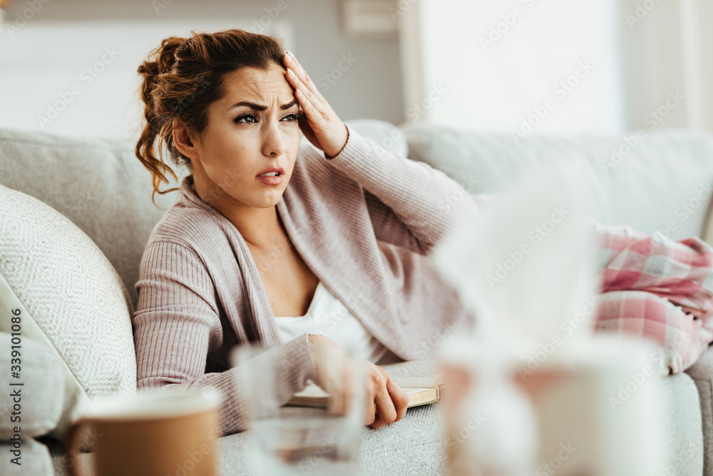 Young woman holding her head in pain while reading book on the sofa.