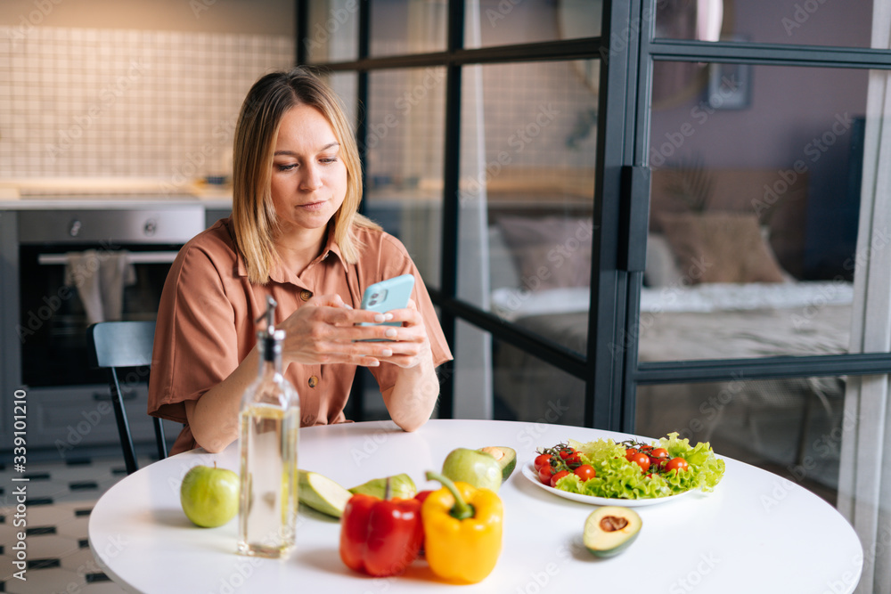 Young woman with fresh vegetables smiling while using phone at the table in kitchen with modern interior. Concept of healthy eating.