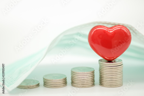 Global Healthcare, Coronavirus,Financial, Covid-19 Protection Concept. Closeup of ceramic red heart on top of stack of coins under sugical mask. photo