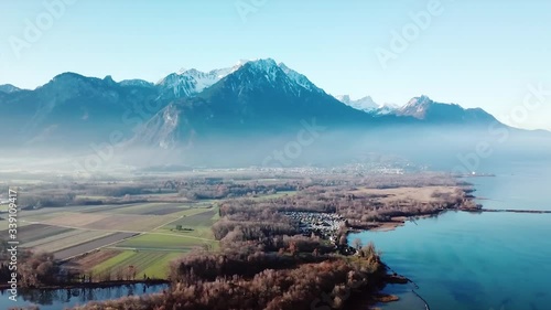Aerial, rising, drone shot panning over calm lake Geneva and frosty forest, mountains in background, foggy morning, in Lausanne, Switzerland