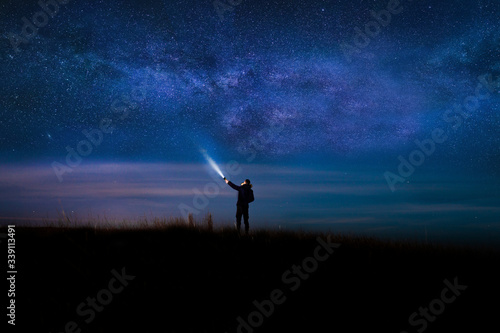 Night sky with  Milky Way and silhouette of a standing happy man with classic blue light. Image soft focus and noise due to long expose and high iso.