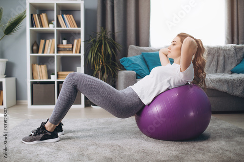 Pretty young woman in activewear doing exercises for abs on pilates ball while staying at home. Athletic girl with blond hair training regularly indoors.