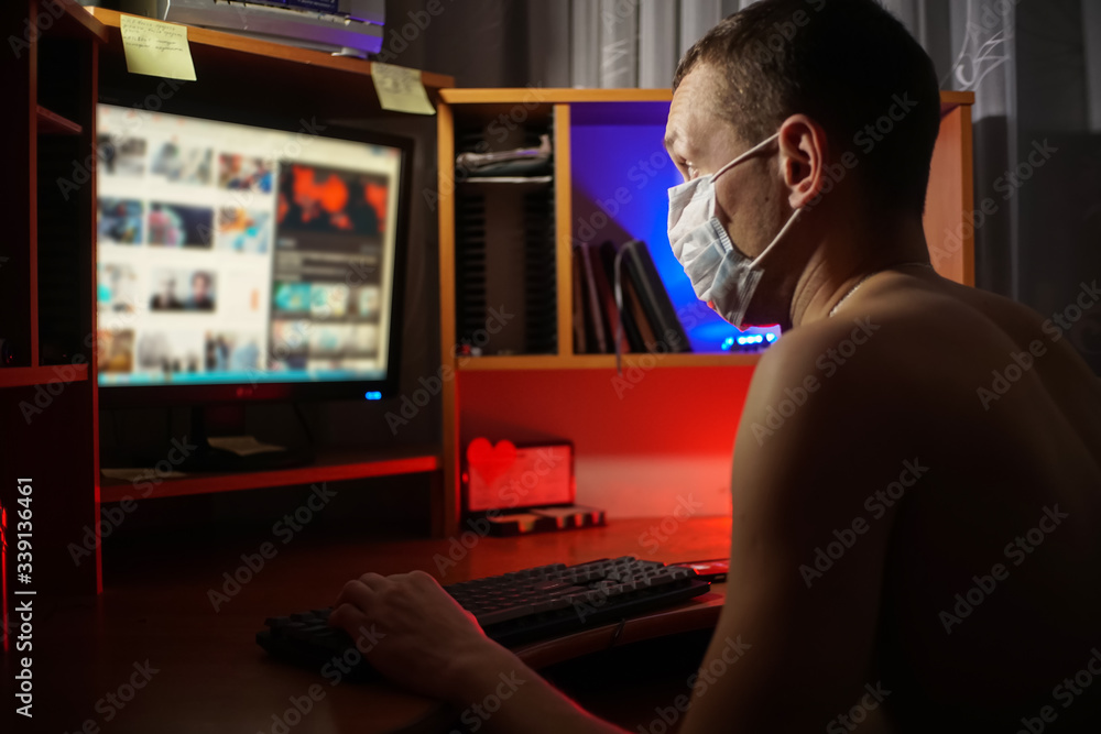 Man with a bare torso in a medical mask sits at a computer and studies information in a dark room. Self-isolation