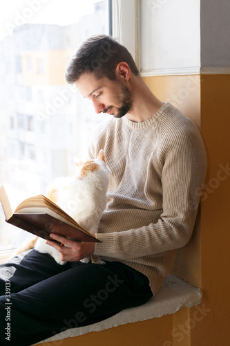 A young man sits on a windowsill and reads a book. Next to the man sits a white cat with red spots. A man is wearing a beige sweater.