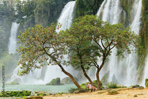 Young female traveler near the big green tree in front of full of water huge waterfall near the green forest with green trees in mountains
