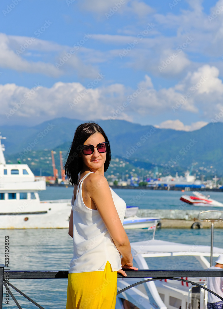 Positive woman walking at the harbor on vacation. Mountains on background. Cheerful, smiling, lucky lady in white blouse, yellow trousers and sunglasses, outdoors at sea port. Sunny summer day.