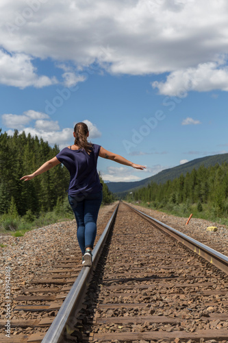 woman from behind balances on the train tracks that run through an incredible landscape between forests and mountains in the Canadian natural parks, on a day with white clouds and blue sky