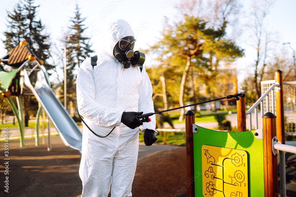 Man wearing protective suit disinfecting the playground in the sun with spray chemicals to preventing the spread of coronavirus, pandemic in quarantine city. Covid -19. Cleaning concept. Man wearing protective suit disinfecting the playground in the sun with spray chemicals to preventing the spread of coronavirus, pandemic in quarantine city. Covid -19. Cleaning concept.