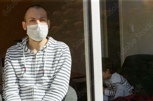 Man wearing face mask sitting by the window and his child during their self-isolation at home as infections and coronavirus disease are spreading. Suffering ill people at home to prevent an epidemic