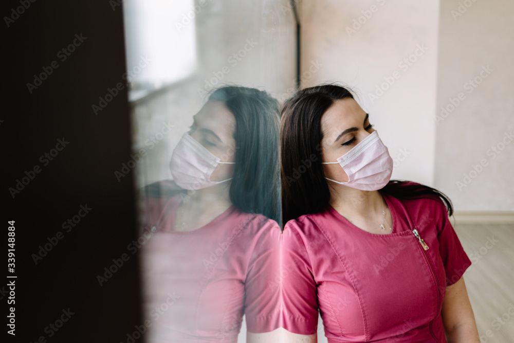 Sad doctor standing near window in hospital face mask because of coronavirus epidemy. Portrait of upset, tired and worried doctor woman thinking of covid-19 pandemic Sad doctor standing near window in hospital face mask because of coronavirus epidemy. Portrait of upset, tired and worried doctor woman thinking of covid-19 pandemic