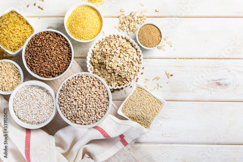 Selection of whole grains in white bowls - rice, oats, buckwheat, bulgur, porridge, barley, quinoa, amaranth, on white wood background