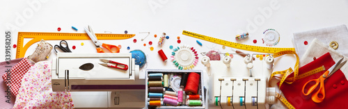 Sewing clothes. Panoramic top view on desktop: sewing machine, box with colored threads, overlock and accessories, scissors, buttons, needles, rulers and fabrics on white background. Banner photo