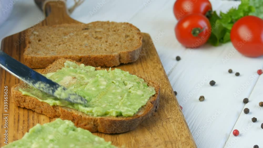 Spread mashed avocado on a toasted bread with a knife. Making tasty avocado toast for breakfast. Cooking bruschetta with cherry tomatoes and quail eggs. Closeup view. Healthy Vegan food. Spread mashed avocado on a toasted bread with a knife. Making tasty avocado toast for breakfast. Cooking bruschetta with cherry tomatoes and quail eggs. Closeup view. Healthy Vegan food.