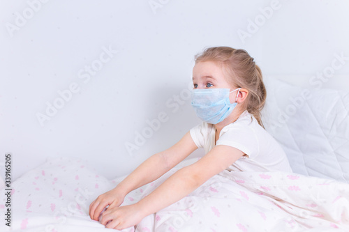 Little blonde girl in protective medical mask lying in white bed in a white room.