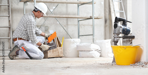 plasterer man construction worker work with tool box wear gloves, hard hat and protection glasses at interior building site with scaffolding. bucket, sacks and jackhammer photo