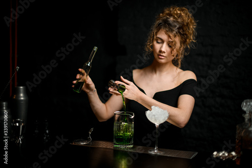 pretty girl bartender in black blouse gently pours drink from jigger into mixing cup.