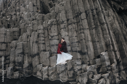 Wedding in Iceland. A young guy and a girl in a white dress and a red sweater are standing on the ledges of the rock